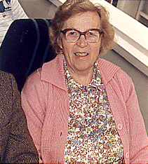 A colour photo of an older woman in a pink cardigan sitting on a chair