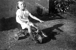 An old black and white photo of a child on a tricycle on the grass outside a house