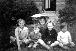 An old black and white photo of four children sitting cross legged on the grass outside a house, with a woman sitting behind them holding a sunshade above her