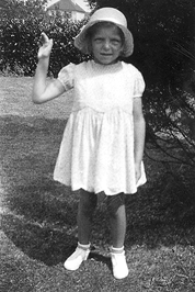 An old black and white photo of a child wearing a sun hat and knee length dress standing in a garden