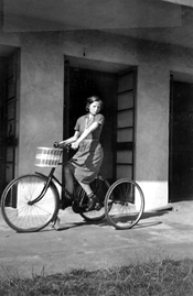 An old black and white photo of a young woman on a bicycle outside a building