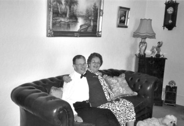 A black and white photo of a man and a woman sitting on a sofa with their arms around each other,  in a homely setting, sith a small white dog at their feet
