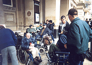 A colour photo showing a group of people, some standing some in wheelchairs, outside a building