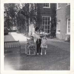 An old black and white photo of a woman outside a manor house, holding a yound child, with another standing beside them and a third child in a wheelchair