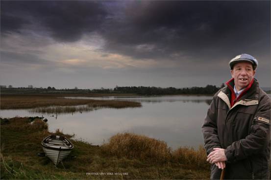 Patrick Kearney standing beside a lake with an old boat on the shoreline