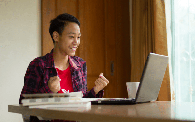 A happy, smiling teenage boy is pictured sitting at a table with a laptop
