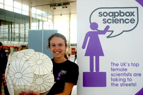 A young female scientist stands next to a Soapbox Science banner stand