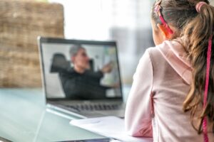 A photo taken from behind of a young woman looking at a person speaking on a laptop screen.