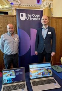 Two men in front of an Open University banner with courses displayed on laptops