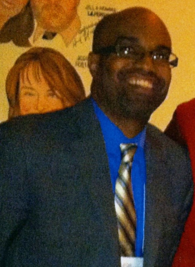 A photo of Ron Buckmire standing in front of a yellow background. Ron is wearing a blue shirt, stripey tie and glasses. He is smiling. 