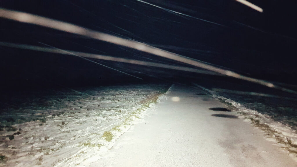 An image taken in the dark with a headtorch of a snow covered path with snow trails blowing across the picture. 