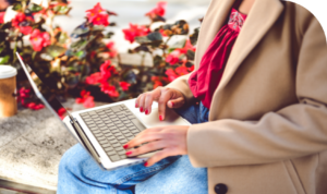 Medium close up photo shot of woman using laptop outside with flowers in the background.