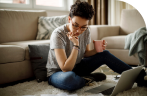 A woman sits on the floor of a living whilst looking calmly at sheets of paper and a laptop. 