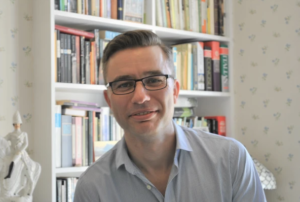 Photograph of James Robson wearing glasses and blue shirt, with books and a classical figurine in the background.