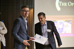 A photograph of a man wearing glasses and a blue jacket. He smiles directly at the camera while holding a paper certificate in his left hand.