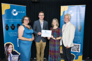 Photograph showing four formally dressed people smiling at the camera. The man and the woman in the centre of the image hold up a certificate.