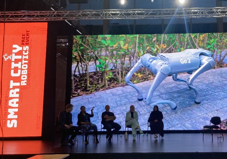 Five human panellists in silhouette against a large screen in which video of a quadrupedal robot is projected. The screen is framed by banners that read "Smart City Robotics" 