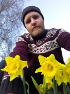 Kevin De Ornellas, a white middle-aged male with a beard. He's wearing a black beanie and a maroon woollen jumper, and he's crouched in front of a patch of daffodils. 