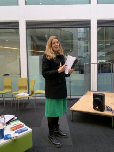 A middle-aged white woman with long blond hair, wearing a green dress. She is standing up and talking. 