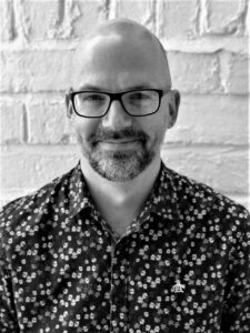 A headshot of a middle-aged bald male with a beard, wearing rectangular shaped glasses and a floral shirt