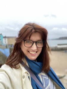 Headshot of a middle-aged woman with brown hair wearing circular glasses, a blue scarf and cream jacket.