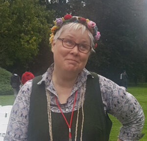 A middle-aged Caucasian woman with short blond hair, wearing long multi-coloured beads and a floral headress. She is looking up to the left-hand corner of the frame, wearing a cheeky grin. 