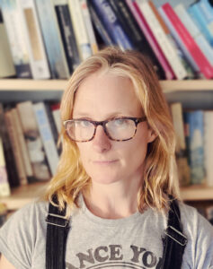 Headshot of middle-aged woman with blonde hair wearing tortoise shell coloured glasses, a grey t-shirt and black dungarees.