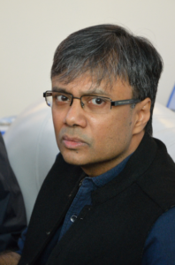 Headshot of an Indian older male, wearing rectangular sunglasses, a blue shirt and a black gilet.