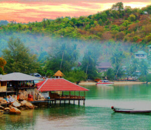 A lake in Thailand, surrounded by lush green hills with a red wooden hut standing on stilts in the water. The sunset and the light reflecting on the water surface creates a picturesque scene of natural Thailand.