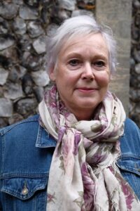 A headshot of a woman with short grey hair, wearing a denim jacket and a white and pastel-pink scarf.