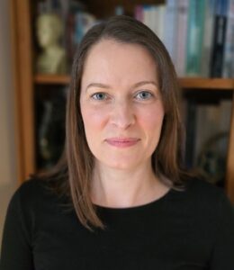 A Caucasian woman with long straight dark hair, standing in front of a bookcase.