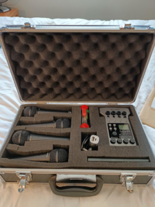 A photograph of a flight case, lined with dark pick-foam, containing four microphones, a range of batteries and a mobile XLR recording interface.