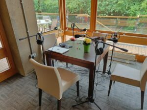 A photograph of a table, with four microphones on boom arms facing four empty chairs. This is at the bank of a glass walled cafe.