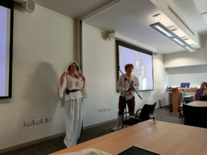 A photograph of Jane Secker and Chris Morrison. They are wearing Star Wars outfits at the front of a conference room, and performing a skit animatedly.