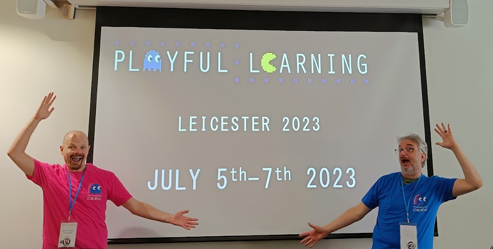 A photograph of Mike Collins and Mark Childs, posing in a silly fashion before a slide showing next years playful learning dates. July 5th-7th 2023. They are having a lovely time.