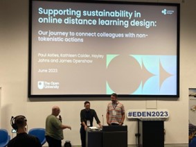Photo of James and Paul speaking at the EDEN conference. They're standing at a podium and there is a large screen behnd them showing slides from the talk.
