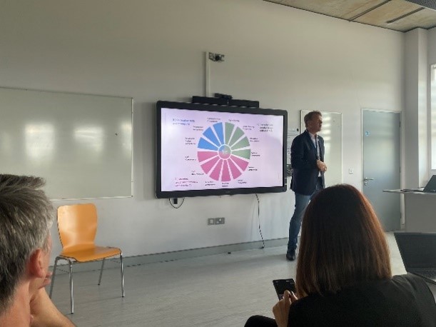 An image of a man giving a presentation. He stands next to a screen that shows a slide which outlines the 12 competencies identified in the presented work related to AI skills for the future. Audience members can be seen in the foreground.