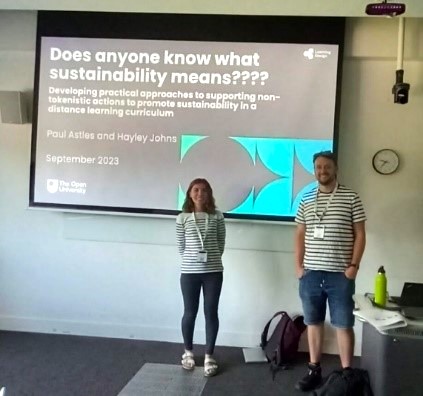 Paul and Hayley next to the screen with the title slide for their presentation. The screen reads: 'Does anyone know what sustainability means???? ; Developing paractical approaches to supporting non-tokenisitic actions to promote sustainability in a distance learning curriculum.'