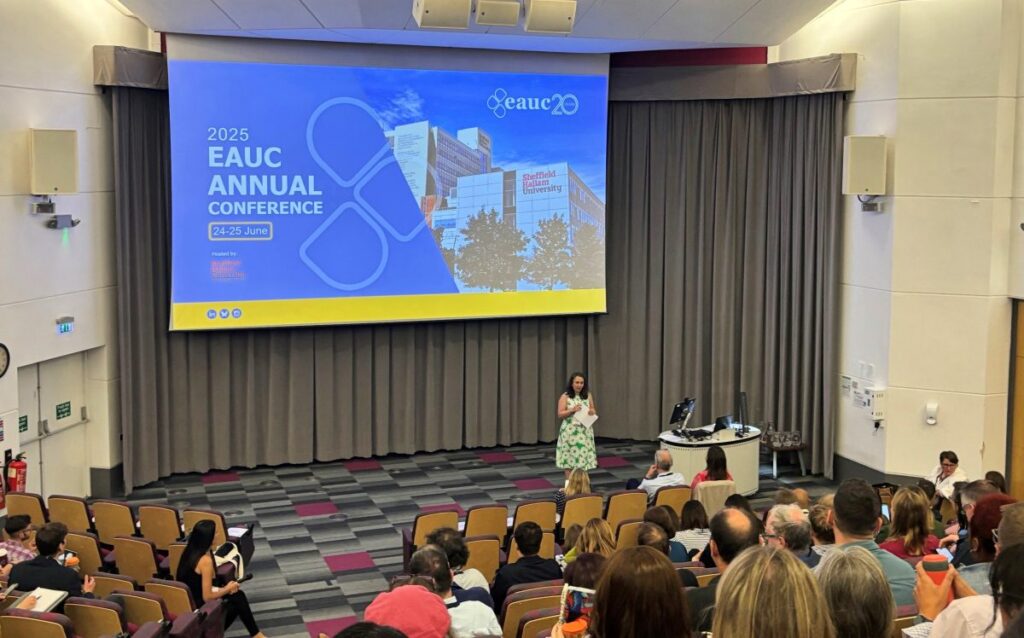 Attendees at the EAUC conference sit in an auditorium. A woman stands at the front presenting. A large screen shows the intro slide to the presentation which reads "2025 EAUC Annual Conference 24-25 June."