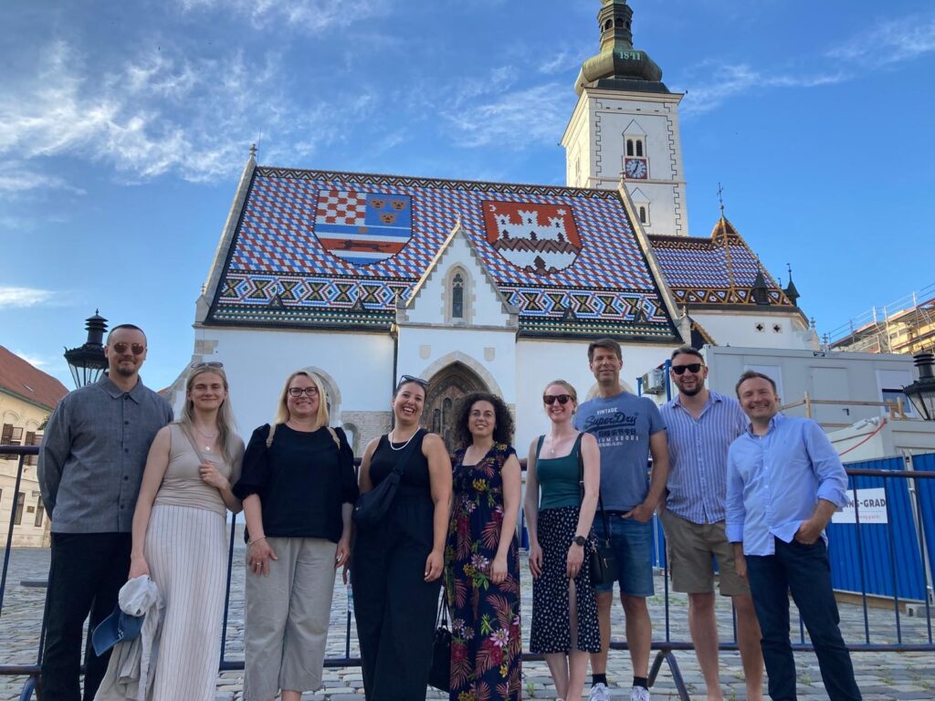 A group of 9 people standing in front of a church with an ornately patterened, tiled roof in Zagreb. Two coats of arms are integrated into the roof tile pattern.