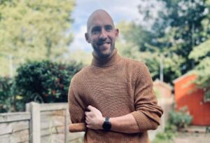 Dan smiling at the camera wearing a brown jumper with trees in the background.
