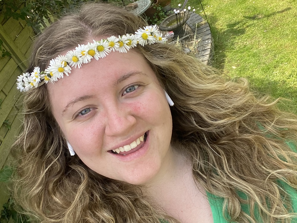 A young women with long blonde hair and a flower crown, smiles at the camera.