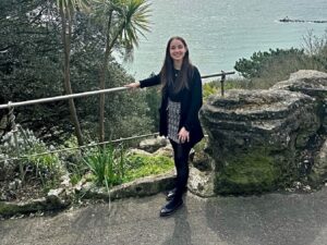 Photo of a young woman with long dark hair, standing in front of a sea view.