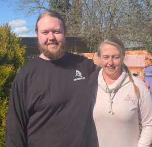 A young man wearing a black top, with his arm around his mum - an older woman with her hair in a plait.