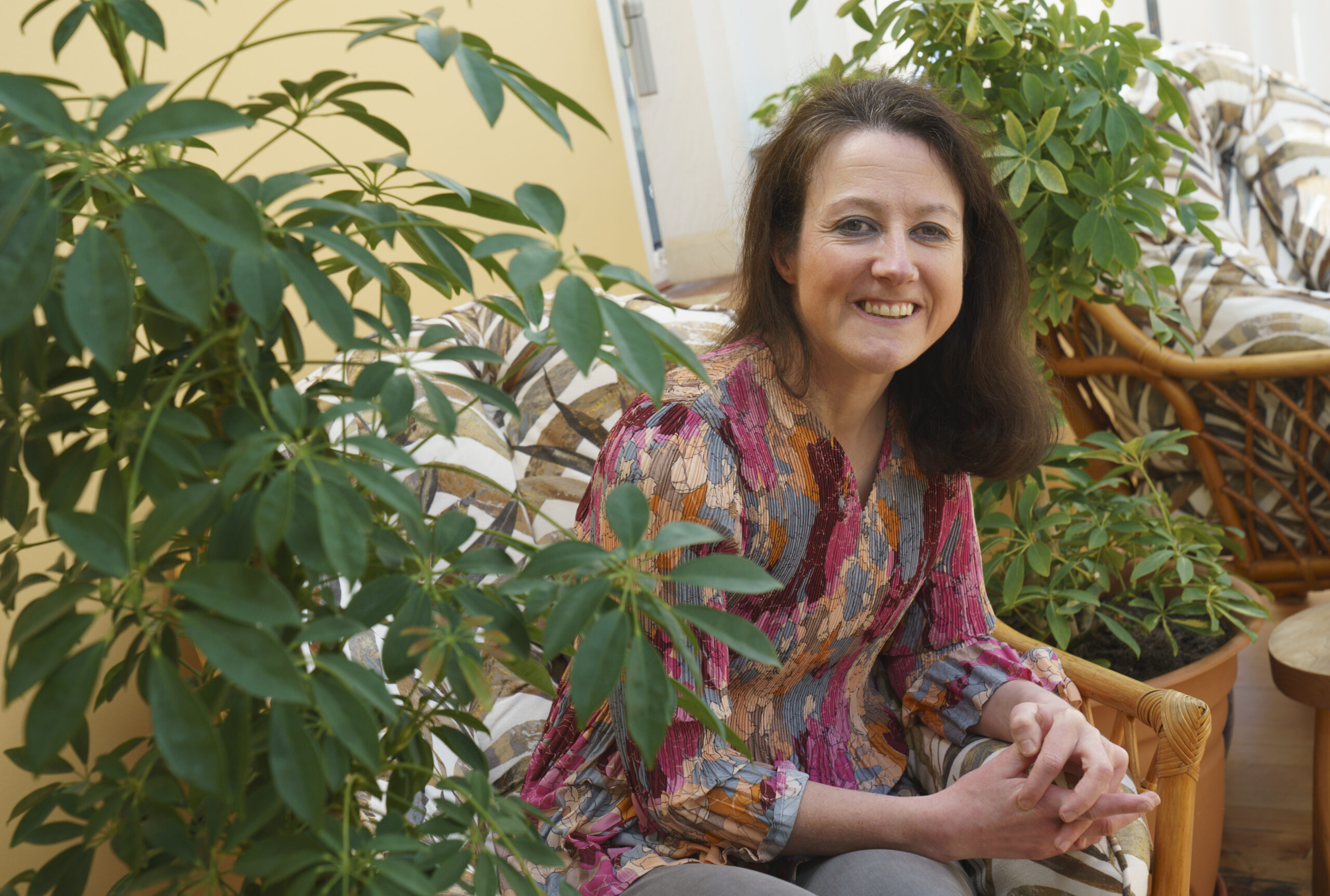 Julie sitting on a chair, smiling at the camera. a leafy plant is on the left of the shot.