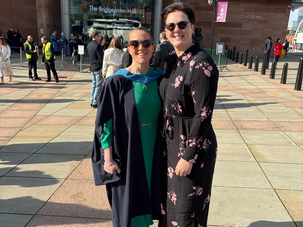 Two women wearing sunglasses, standing next to each other. The woman on the left is wearing graduation robes.