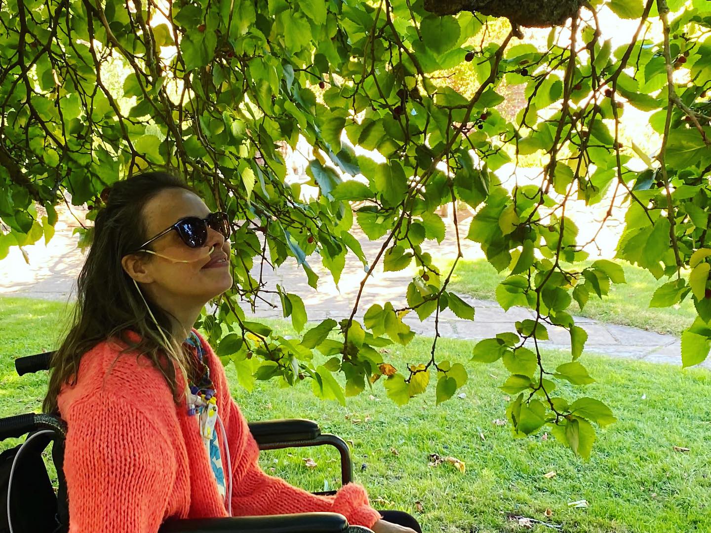 Jade sitting in a wheelchair wearing sunglasses, under a tree.