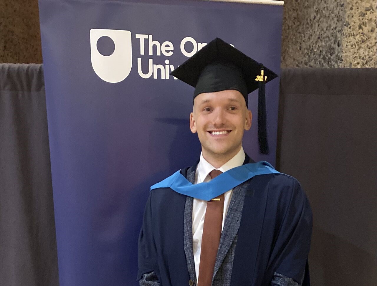 JAke standing in front of an Open University banner, smiling, wearing graduation cap and gown.