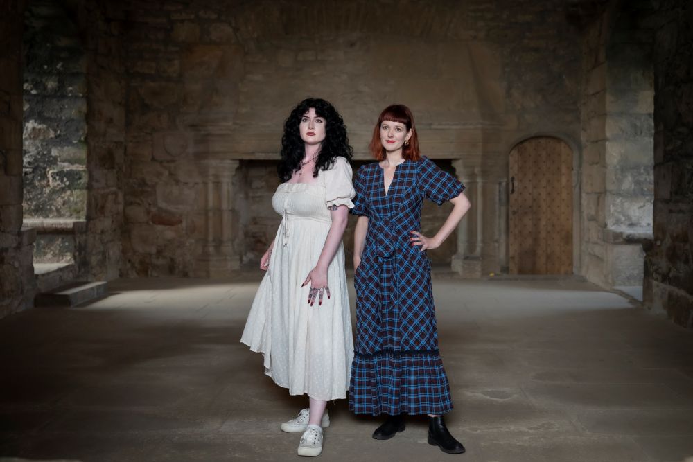 Presenters Evelyn in a white dress and Zara in blue dress standing in a castle setting. Credit: Rogan Productions