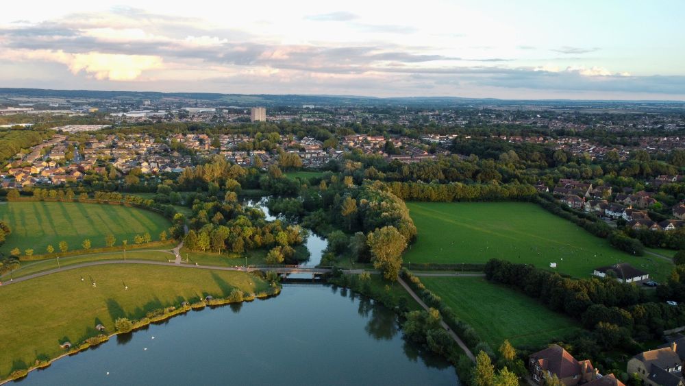 Aerial view of Milton Keynes, showing green parkland, lakes, tree-lined paths and surrounding residential areas stretching towards the horizon.